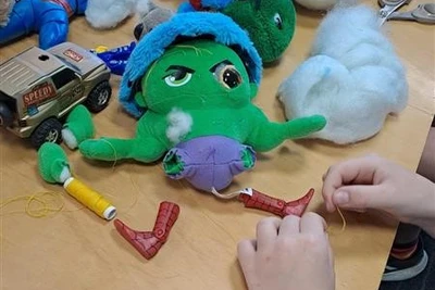 A green stuffed toy with its legs removed. A child's hands are sizing up some spiderman legs to replace the toy's legs