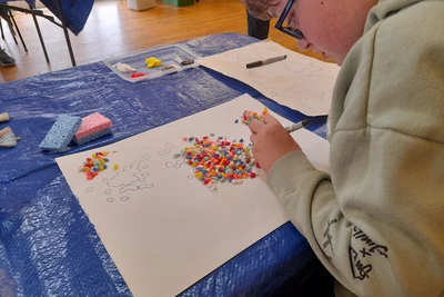 Small gravel is spread across a sheet of paper, and a child draws around it to create the outline of an island.