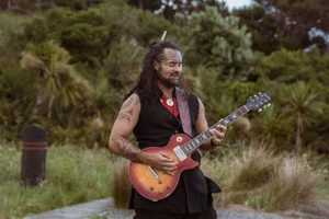 Matiu Te Huki stands in front of native bush playing a guitar