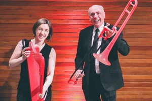 A woman in a black dress holding a red harp stands next to a man in a suit holding a red trombone, both are smiling.