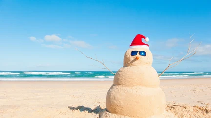 Beach snow man made of sand against a blue summery sky, with sunglasses and Santa hat on