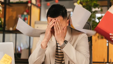 Photo of a person at a work desk with head in hands surrounded by flying papers.