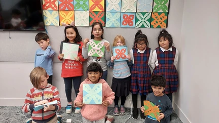 A group of children are standing in front of a display of traditional Sāmaon siapo (tapa) cloths, proudly displaying paper versions that they have made.