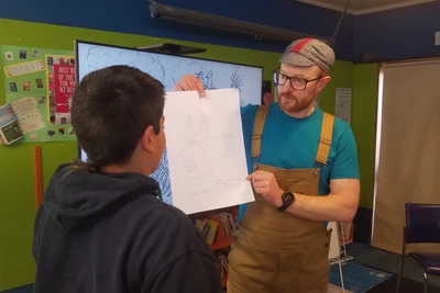 Steve, wearing a small cap and brown overalls over a blue tshirt holds up a participant's design, while the young inventor explains it to the group.