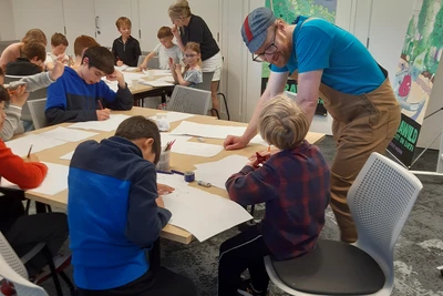 Five children are working on designs at a table covered in sheets of paper. Steve leans over as one participant shows him their design.