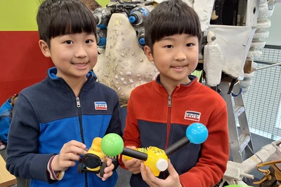 Two boys hold a small yellow puppet creature with one blue and one green antenna.