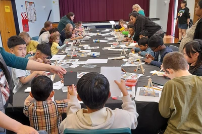 A long table in the middle of a hall surrounded by children painting tiny artworks.