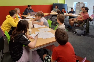 A young man sites behind a card DM screen in front of a group of young people seated at tables.