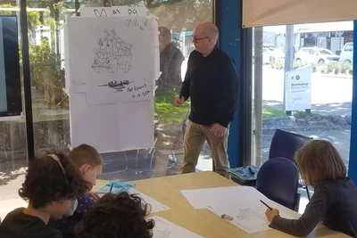 Paul Beavis gestures at a drawing of Nee-naw the fire engine in front of an engaged group of children.