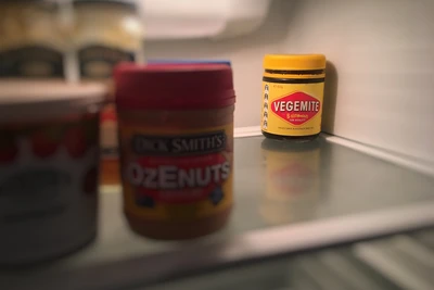 An unopened jar of vegemite sits at the back of an open fridge.