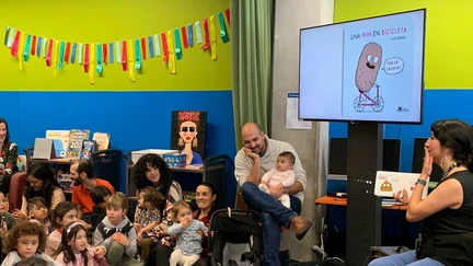 A librarian reads a book in Spanish to a large group of children and parents. A screen is displaying illustrations from the book with accompanying captions in Spanish.