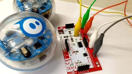 Two Spheros (spherical robots with transparent external panelling so you can see the circuitry inside) sit next to a Makey Makey kit, a programmable microcomputer that can be connected to real-world objects using alligator clips.