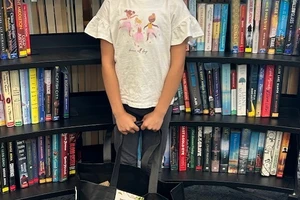 A smiling girl with a pink headband stands in front of a library bookshelf holding a large bag of prizes