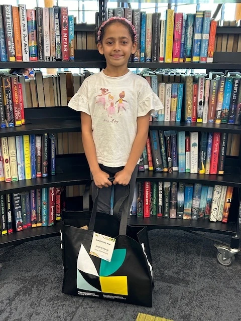 A smiling girl with a pink headband stands in front of a library bookshelf holding a large bag of prizes