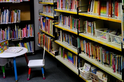 Yellow edged bookshelves in the corner of the library. In the foreground is a little table and chair with giant pencils in place of legs.