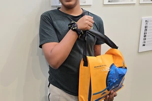 A grinning boy stands in front of a display of black and white photos holding an orange library satchel full of prizes.