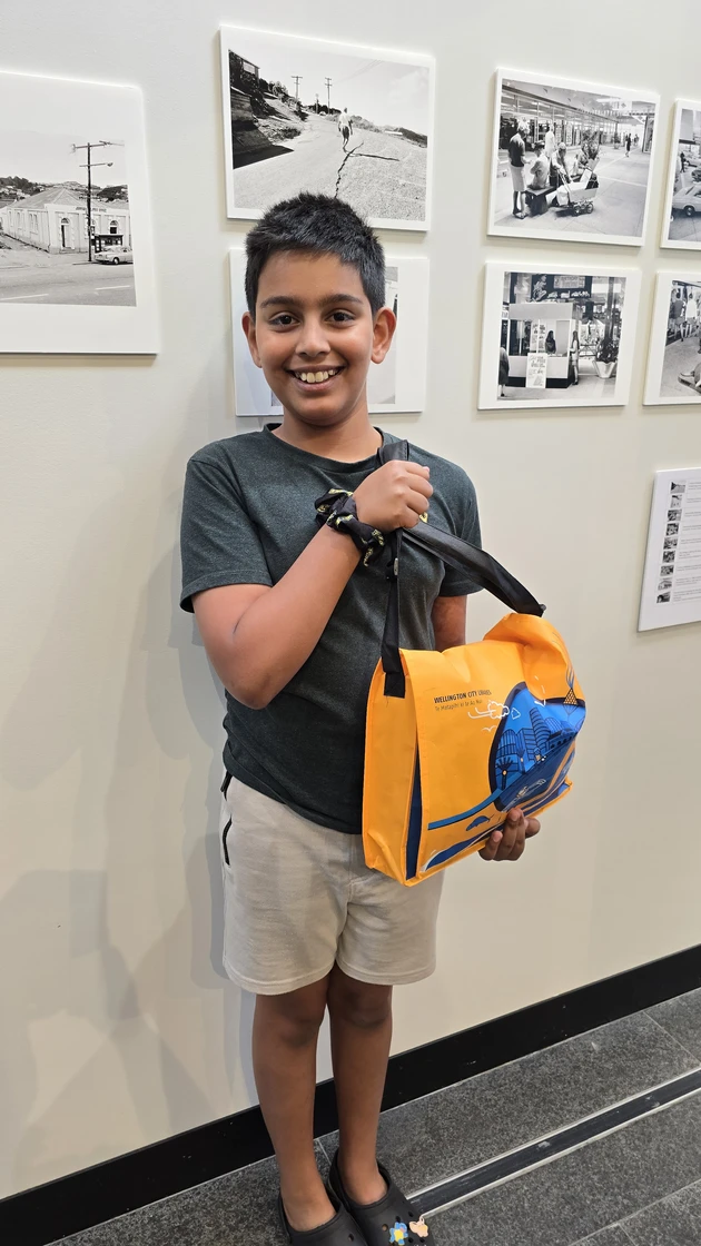 A grinning boy stands in front of a display of black and white photos holding an orange library satchel full of prizes.