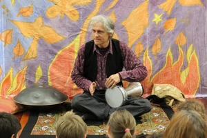 A man sitting cross-legged on a mat in front of a crowd of children, raising his hands to play a drum