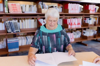 Older māori wahine reading a document at a table in a library, with bookshelves in the background.