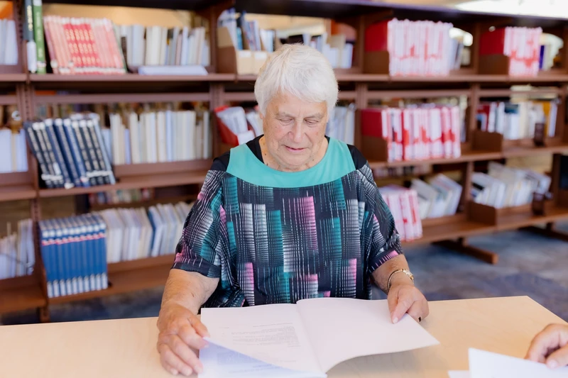 Older māori wahine reading a document at a table in a library, with bookshelves in the background.