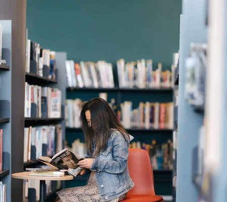 Person sitting on a chair in a library aisle, reading a book with shelves of books around them.