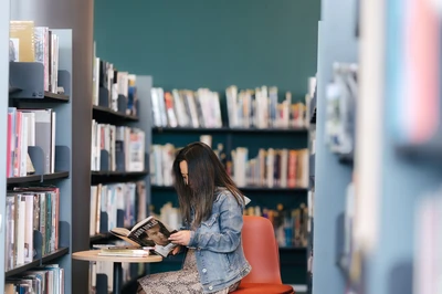 Person sitting on a chair in a library aisle, reading a book with shelves of books around them.