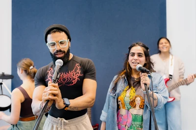 People singing into microphones in a recording studio, with others playing instruments in the background.