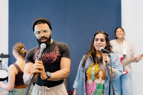 People singing into microphones in a recording studio, with others playing instruments in the background.