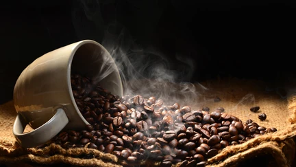 An overturned coffee cup on a pile of coffee beans with steam rising from it