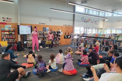 Josh, wearing pink overalls and a beanie with a pig face on it raps into a microphone. Sacha, in a sparkly fringed pink jacket, dances next to him