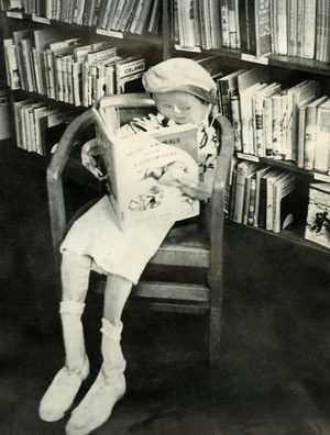 B&W image of a deflated child, definitely not a child-sized doll, seated on a chair in a library, holding a book in their hands.