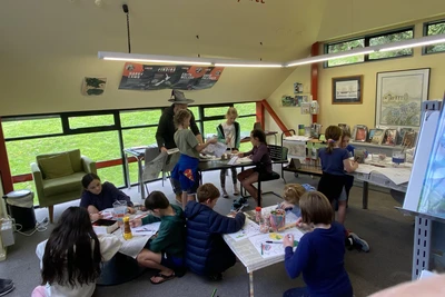 Four busy tables of children drawing maps, in the background a librarian with a wizard's hat helps lay out a table.