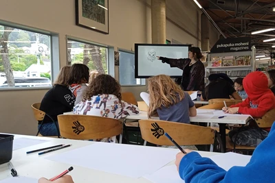 In front of a seated group if children, Izzy points to an illustration of a taniwha face