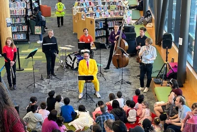 Chris Sam Lam in a bright yellow suit sits in front of a flautist, trumpet player, percussionist, double bass player, and viola player