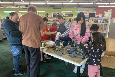 Parents and children gather around a table covered with power drills, other tools, and recycled items to use to make puppets.