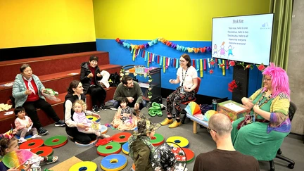 Two librarians, both wearing colourful costumes and wigs, are reading stories to a group of young children and their families, all similarly wearing bright costumes and hats.