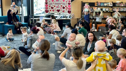 A large group of parents and caregivers with babies sit on cushions and chairs, all facing towards a librarian who is holding a baby doll and singing a song. Many are lifting their babies high into the air.