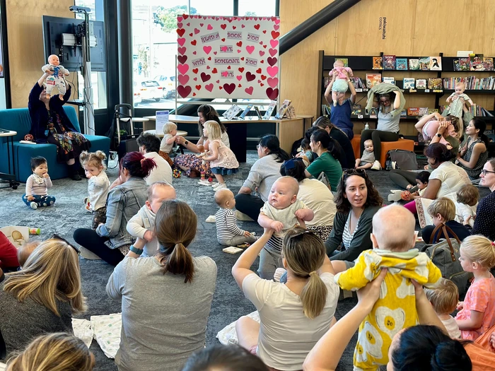 A large group of parents and caregivers with babies sit on cushions and chairs.