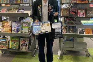 A smiling teen in school uniform with both her lapels covered in badges, holds a library bag of prizes, a book, two certificate