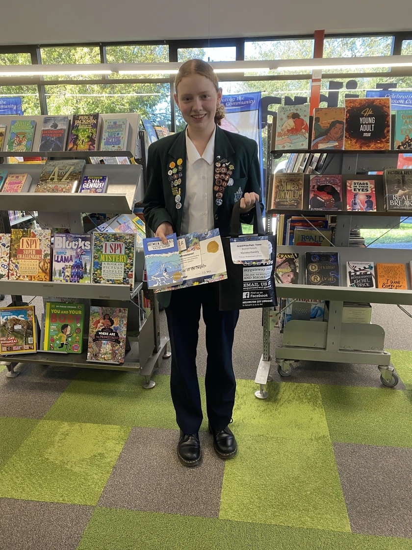 A smiling teen in school uniform with both her lapels covered in badges, holds a library bag of prizes, a book, two certificate