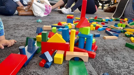 A small pile of colourful blocks lies on the floor. Some have been arranged into some kind of structure. Children play in the background.