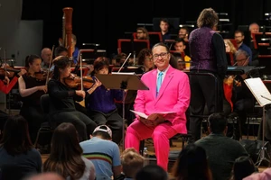 A man in a vibrant pink suit sitting in front of a group of violin players and a harpist talks to an audience of children and parents