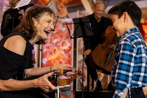 A smiling violin player bends down to show a child her violin