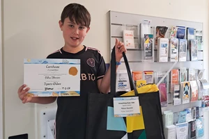 A boy smiles, holding a certificate in one hand and a bag of prizes in the other