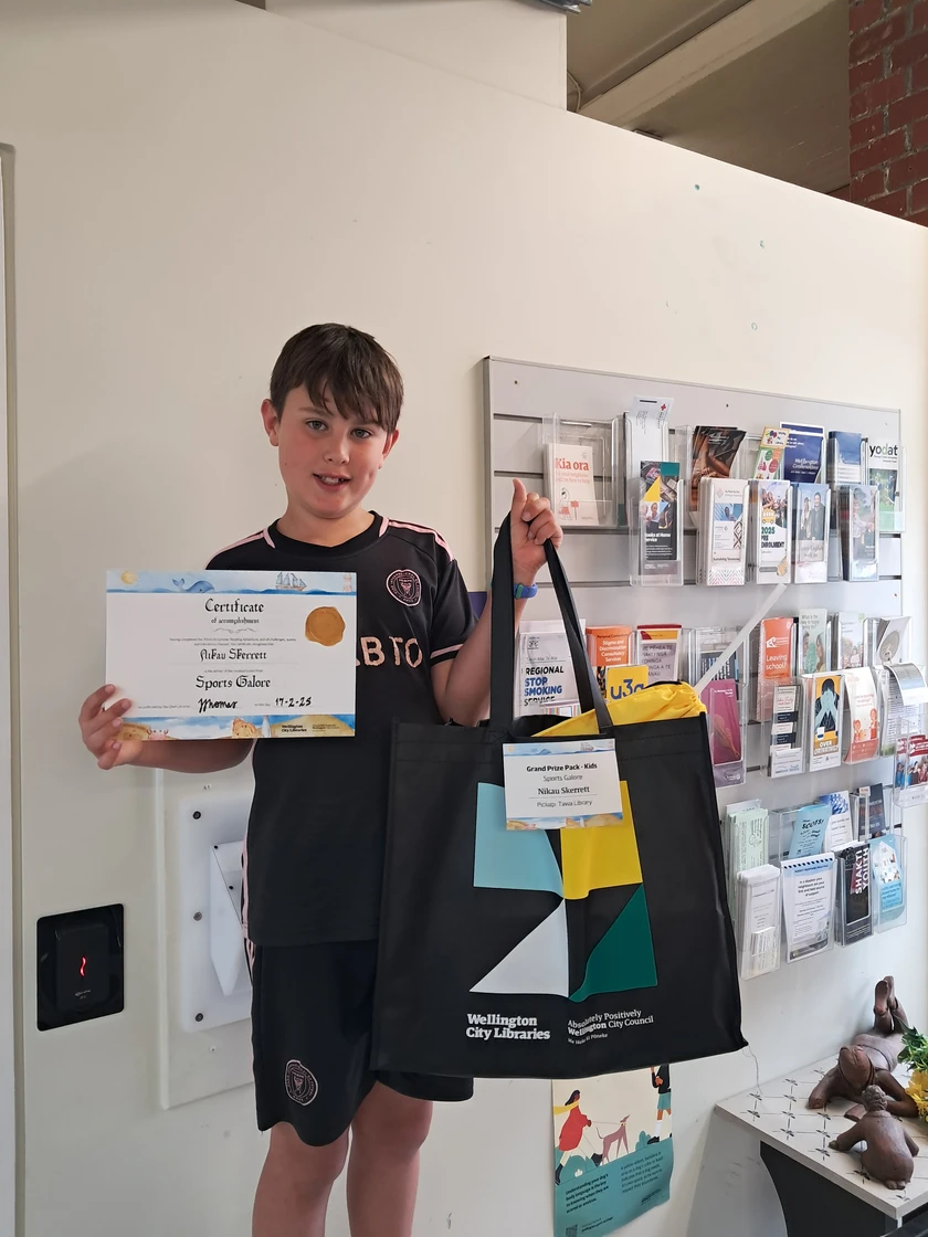 A boy smiles, holding a certificate in one hand and a bag of prizes in the other