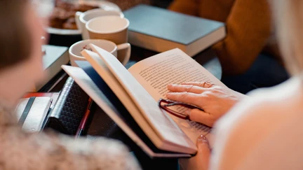 People sharing a book, with a cup of tea and plate of biscuits visible on the table in front