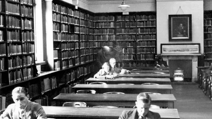 Black and white photograph, lines of tables and bookcases lining the walls. Someone at a table is lit up by light coming through a window, behind them are two blurry figures