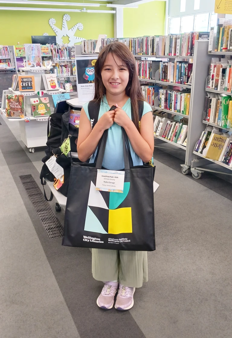 A smiling girl holds a large library tote bag full of prizes up to her chest