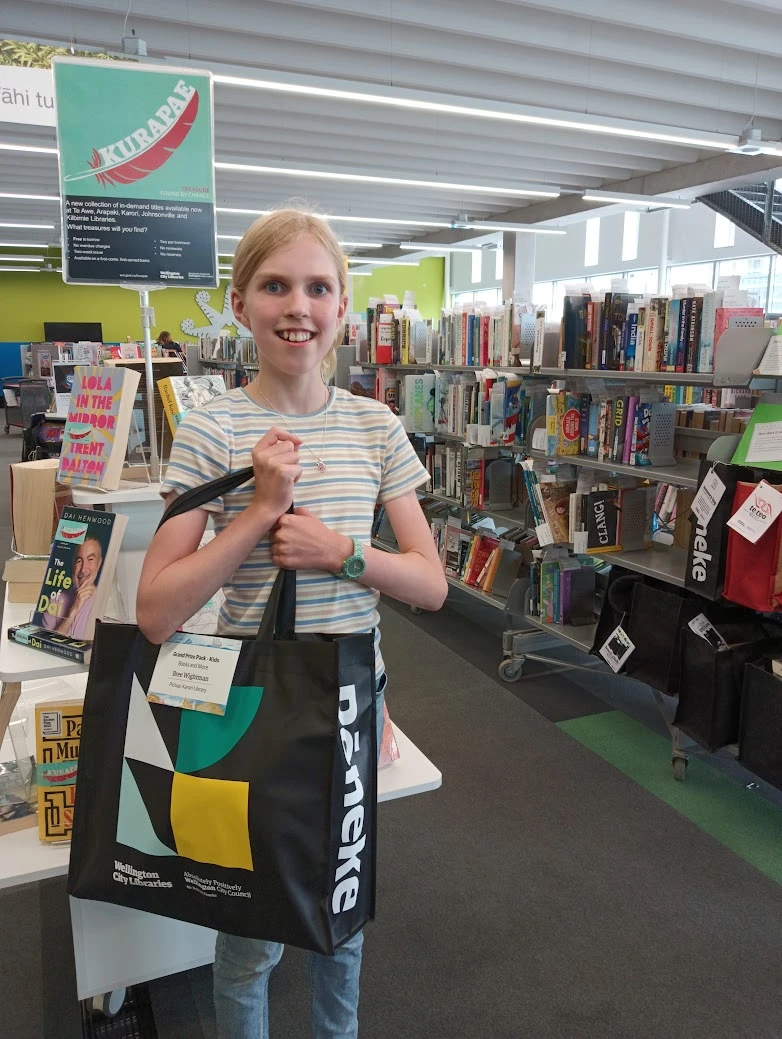 A girl in a stripy shirt grins while holding a large library bag full of books