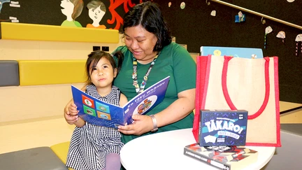 A woman sits with her child, reading aloud from a children's book in te reo Māori, with a small pile of te reo books and one of the library's Te Reo Kete next to them.